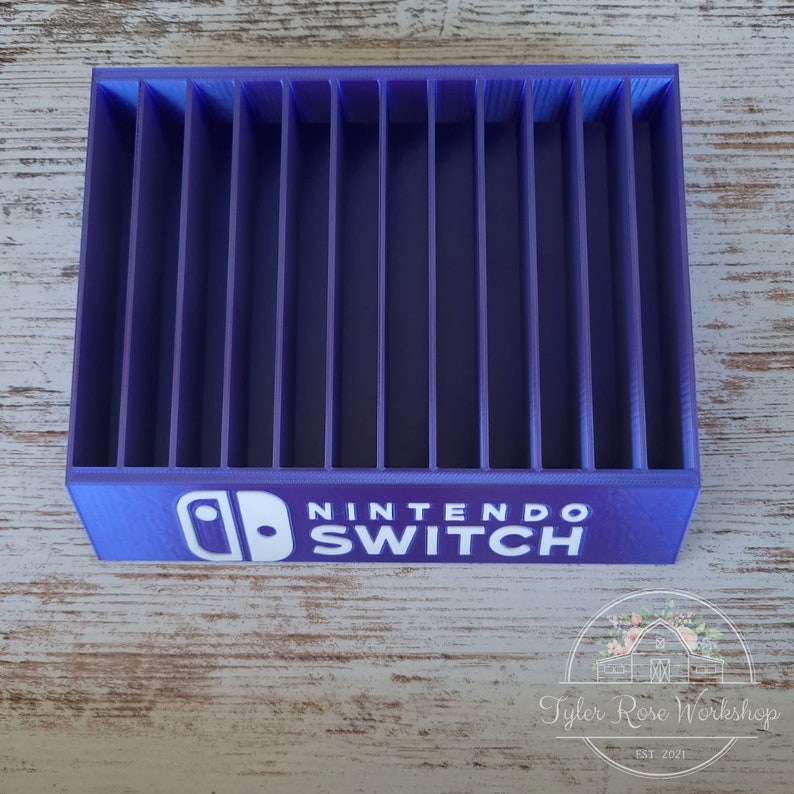 May include: A purple Nintendo Switch game cartridge organizer with vertical dividers. The Nintendo Switch logo and text are printed in white on the front. The organizer is made of plastic and sits on a light wood surface.