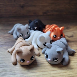 Pode incluir: Um grupo de seis pequenas estatuetas de gato enroladas em várias cores, incluindo cinza, branco, laranja e marrom. As estatuetas têm olhos pretos e narizes cor de rosa.
