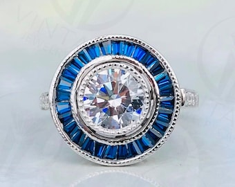 May include: A silver ring with a large, round, clear center stone surrounded by a halo of small, rectangular, blue stones. The ring has a detailed, vintage-inspired design, with a textured band and intricate detailing around the stones.
