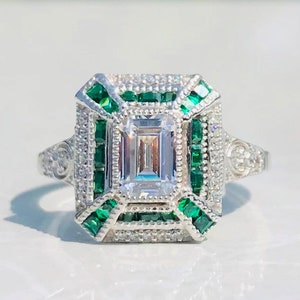 May include: A silver ring with a square-shaped center stone surrounded by smaller stones. The center stone is a clear emerald cut diamond, and the surrounding stones are green emeralds.