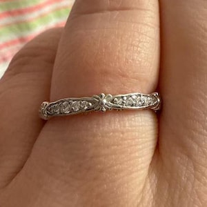May include: A silver ring with a floral design and a row of small, clear gemstones. The ring is set against a blurred background.