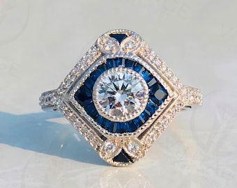 May include: A silver ring with a large round center stone surrounded by a halo of smaller stones. The halo is set with blue sapphires and the ring has a square-shaped setting.