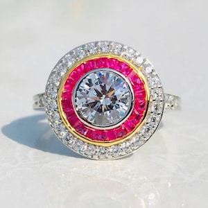 May include: A silver ring with a large, clear, round center stone. The ring features a vibrant pink band surrounded by a gold band and a halo of smaller, clear stones. The ring is set on a white surface.