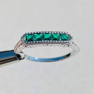 May include: A silver ring featuring five square-cut emerald green gemstones set in a rectangular design. The ring has an ornate, filigree-style band with delicate detailing. The gemstones are a vibrant green color.
