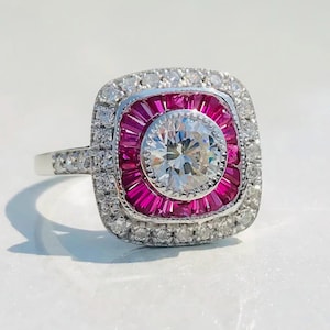 May include: A silver ring with a square face. The center features a large, clear, round-cut stone surrounded by a ring of small, rectangular, pink stones. The outer edge is lined with small, clear stones.