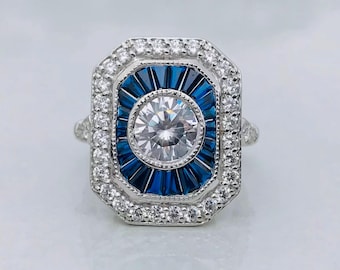 May include: A silver ring with a rectangular face. The ring features a central, round, clear stone surrounded by a halo of blue stones. The outer edge is lined with small, clear stones. The ring has a vintage, art deco style.