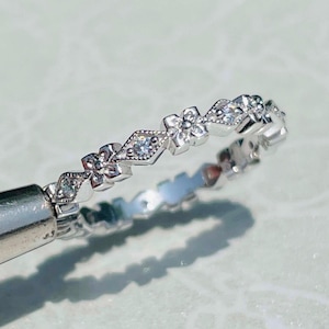 May include: A silver-toned ring featuring a repeating pattern of small four-petaled flowers and diamond-shaped settings. Each flower and diamond shape contains a small, clear, sparkling stone. The band has a delicate, intricate metalwork design.