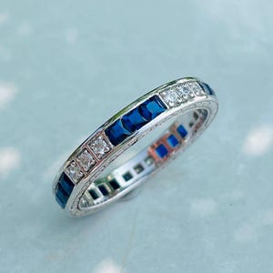 May include: A silver ring with alternating square-cut blue gemstones and clear square-cut gemstones. The ring has an engraved design along the band. The ring is set against a light blue background.