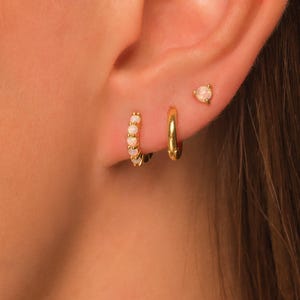 May include: Close-up of an ear adorned with three gold earrings. One is a small stud with a white gemstone, and the other two are hoop earrings. One hoop is a small, plain gold design, and the other is a gold hoop with white gemstones.