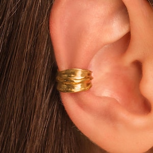 May include: A gold ear cuff featuring a wide band with a textured, organic, and wavy surface. The metal has a polished finish that reflects light, creating highlights and shadows across the uneven, rippled contours of the circular jewelry piece.