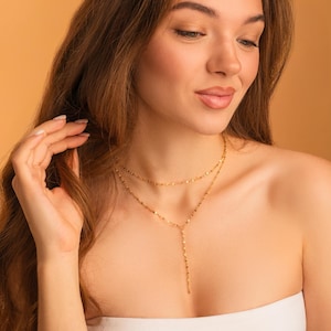 May include: A gold-toned layered necklace featuring two delicate chains with small, reflective, flat metallic accents. The upper chain sits as a choker, while the lower chain includes a vertical drop segment that hangs down from the centre point.