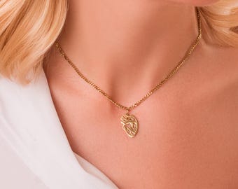 May include: Gold-coloured necklace with a pendant featuring an abstract heart design. The pendant hangs from a delicate chain. The necklace is worn against a white shirt.