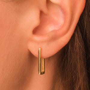 May include: A close-up of a gold-colored rectangular earring. The earring is a simple, geometric design, with a vertical bar shape. The earring is worn on a person's ear.