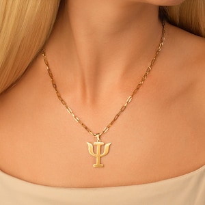 May include: Gold-coloured necklace with a Psi symbol pendant. The pendant is suspended from a delicate chain. The necklace is shown on a person's chest, with the chain and pendant clearly visible.