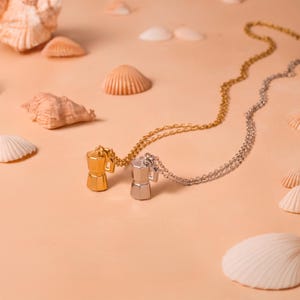 May include: Two necklaces featuring coffee pot pendants. One is gold-coloured, the other silver. They rest on a light orange surface with seashells. The chains are delicate, and the pendants are detailed, evoking a coffee theme.