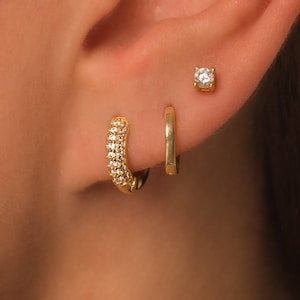 May include: Close-up of an ear adorned with three gold earrings. A small, square-cut stud earring with a clear stone sits at the top. Below, a small gold hoop and a larger gold hoop with small, clear stones are visible.