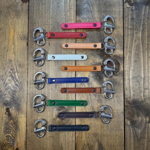 May include: A collection of colorful leather keychains with metal clasps. The keychains are in various colors, including pink, red, tan, light blue, orange, blue, brown, green, and black. They are arranged on a wooden surface.