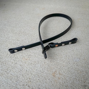 May include: A black strap made of a smooth, flexible material, featuring a looped design secured with a metal buckle. The strap includes multiple silver-toned metal rivets along its length, providing adjustable attachment points for various uses.