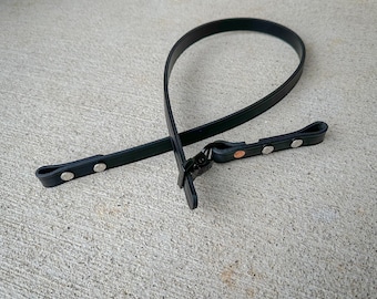 Puede incluir: Una correa negra hecha de un material liso y flexible, con un diseño en bucle asegurado con una hebilla de metal. La correa incluye múltiples remaches metálicos de tono plateado a lo largo de su longitud para diversos puntos de sujeción.