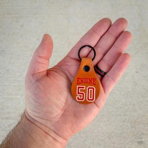 May include: A teardrop-shaped tan leather keyring featuring a black metal ring and a black rivet. The leather is embossed with the red text ENGINE and the number 50 in white with a red outline, centred on the surface of the keyring.