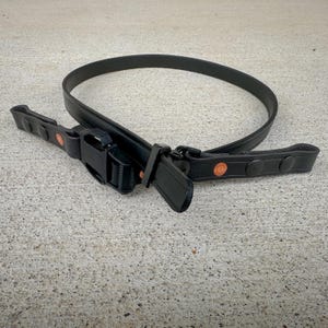 May include: A black belt featuring a durable plastic side-release buckle and adjustable strap. The belt includes two black loops with copper-colored rivets on each side, providing a secure fit and a functional design for everyday utility and wear.
