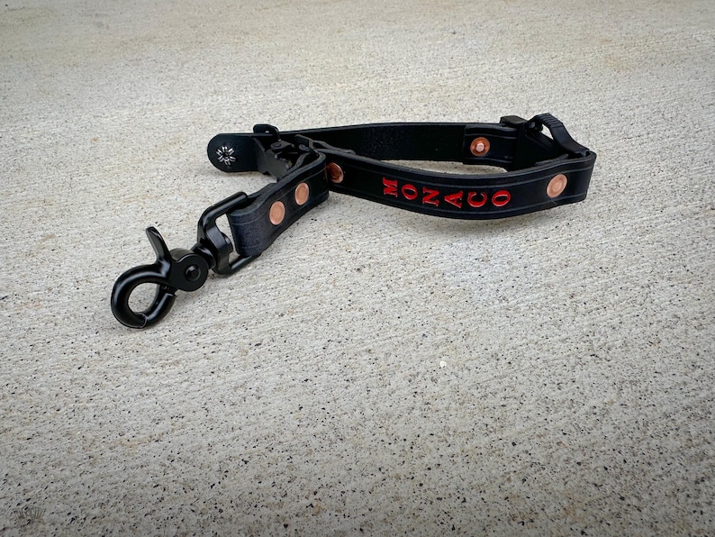 Puede incluir: Una correa negra con acabado mate que presenta un mosquet&oacute;n giratorio de metal, remaches de color cobre y la palabra MONACO grabada en letras rojas. La correa est&aacute; construida con un mecanismo de hebilla e incluye un peque&ntilde;o detalle de logo.