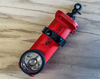 May include: A bright red right-angle torch holder featuring a black leather strap with two black snaps, a black metal swivel clip at the top, and a circular lens opening. The text "SURVIVOR" is visible on the side. Text reads: FLASHLIGHT NOT INCLUDED.