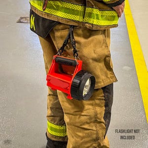 May include: A bright red plastic flashlight holder with a black handle and a black circular lens housing. The holder is attached to a tan firefighter turnout coat with yellow reflective trim using two black metal clips and a Y-shaped strap.