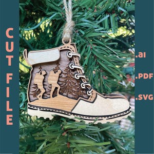 Puede incluir: Adorno navideño de madera con forma de bota de senderismo. La bota presenta un diseño recortado de excursionistas y árboles. El adorno cuelga de un lazo de cuerda. El texto "CUT FILE" está en el lado izquierdo de la imagen.