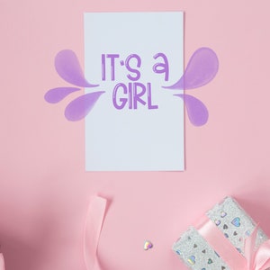 Pu&ograve; includere: Un biglietto rosa con il testo "It's a girl" in lettere viola. Il biglietto &egrave; circondato da forme viola simili ad acquerello. Due scatole regalo con nastro rosa sono sullo sfondo rosa.