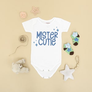 Pu&ograve; includere: Un body bianco per neonato con testo blu che dice "Mister Cutie" con piccole stelle blu intorno al testo. Il body &egrave; su uno sfondo marrone chiaro con un peluche, una stella di legno e una palla di spago.