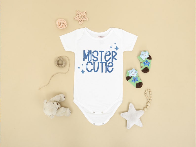 Pu&ograve; includere: Un body bianco per neonato con testo blu che dice "Mister Cutie" con piccole stelle blu intorno al testo. Il body &egrave; su uno sfondo marrone chiaro con un peluche, una stella di legno e una palla di spago.