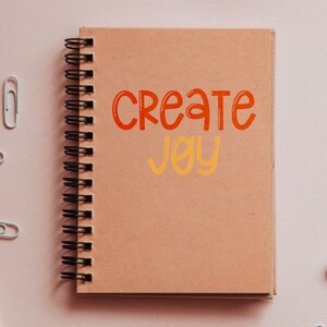 Pu&ograve; includere: Un quaderno a spirale marrone con le parole "Create Joy" scritte in arancione e giallo sulla copertina.
