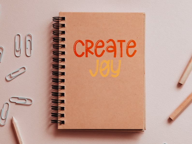 Pu&ograve; includere: Un quaderno a spirale marrone con le parole "Create Joy" scritte in arancione e giallo sulla copertina.