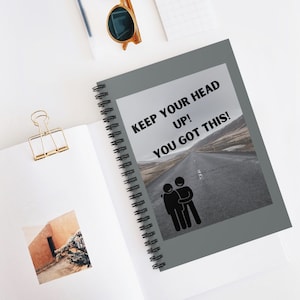 May include: A spiral-bound notebook with a gray cover featuring the text "KEEP YOUR HEAD UP! YOU GOT THIS!" above an image of a road and two stick figures embracing. The notebook is open on a white surface.