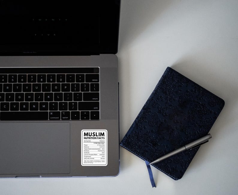 May include: A silver laptop with a rectangular sticker featuring a black and white nutrition facts label titled MUSLIM NUTRITION FACTS, listing values for prayer, patience, kindness, and forgiveness, placed next to a dark blue textured notebook and pen.