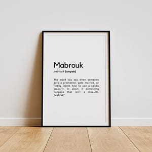 Peut inclure: Une impression encadrée en noir avec le mot "Mabrouk" en gras, suivi de l'orthographe phonétique "mab-rou-k" et de la définition "[congrats]". Le texte se poursuit par une description de la signification et de l'utilisation du mot dans la culture arabe.