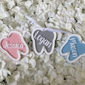 May include: Three tooth-shaped ornaments with white outlines and names written in white. The tooth on the left is pink with the name "Jessica" written inside. The tooth in the middle is gray with the name "Logan" written inside. The tooth on the right is blue with the name "Mason" written inside.