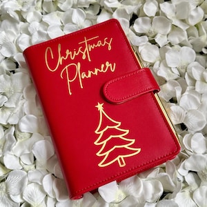 Puede incluir: Planificador de cuero rojo con un &aacute;rbol de Navidad dorado y las palabras "Christmas Planner" en escritura dorada.