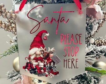May include: A white sign with a red border and red text that says "Santa Please Stop Here". A cartoon gnome and reindeer are holding a stack of presents with a tag that says "Santa's Little Helper".