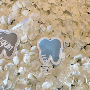 May include: Two white tooth-shaped ornaments with the names "Logan" and "Mason" written on them. The ornaments are on a white background.
