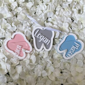 May include: Three tooth-shaped ornaments with white frames and different colored backgrounds. The names "Jessica", "Logan", and "Madison" are written in white on the ornaments.