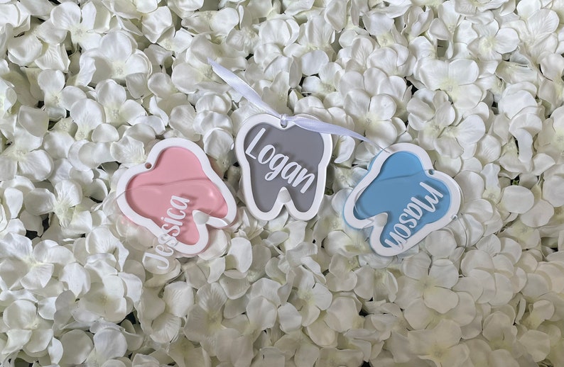 May include: Three tooth-shaped ornaments with white frames and different colored backgrounds. The names "Jessica", "Logan", and "Madison" are written in white on the ornaments.