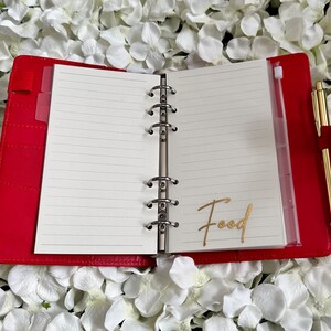 Puede incluir: Planificador encuadernado en cuero rojo con anillas doradas y un bolsillo de pl&aacute;stico transparente en el lado derecho. El planificador est&aacute; abierto en una p&aacute;gina en blanco con la palabra "Food" escrita en letras doradas. Un bol&iacute;grafo dorado est&aacute; en el bolsillo.