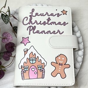 May include: White Christmas planner with a gingerbread house and gingerbread man design. The planner has the text "Laura's Christmas Planner" in pink lettering.