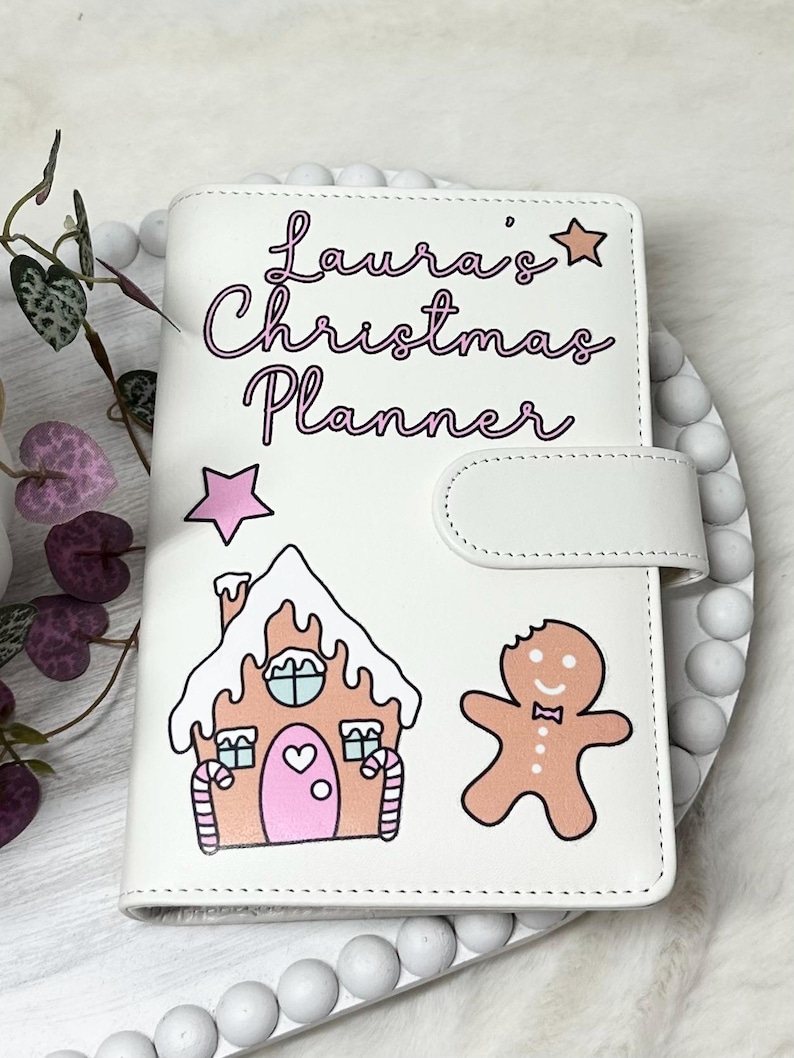 May include: White Christmas planner with a gingerbread house and gingerbread man design. The planner has the text "Laura's Christmas Planner" in pink lettering.