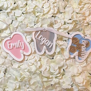 May include: A tooth-shaped banner with three sections, each with a different name. The first section is pink and says "Emily", the second is gray and says "Logan", and the third is blue and says "Bradley".