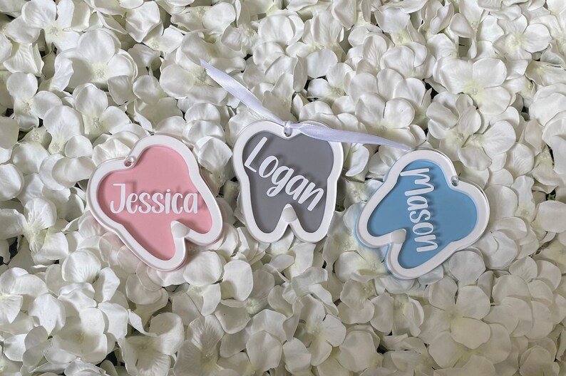 May include: Three tooth-shaped ornaments with white outlines and names written in white. The tooth on the left is pink with the name "Jessica" written inside. The tooth in the middle is gray with the name "Logan" written inside. The tooth on the right is blue with the name "Mason" written inside.