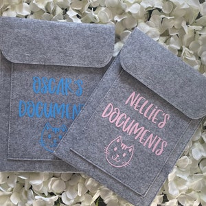 May include: Two grey felt document holders with a pocket on the front. One holder has the text "Oscar's Documents" in blue with a blue cat graphic. The other holder has the text "Nellie's Documents" in pink with a pink cat graphic.