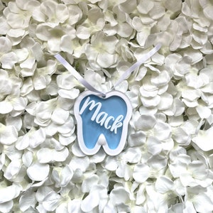 May include: A blue tooth-shaped ornament with the name "Mack" written in white on it. The ornament is hanging from a white ribbon and is surrounded by white flowers.